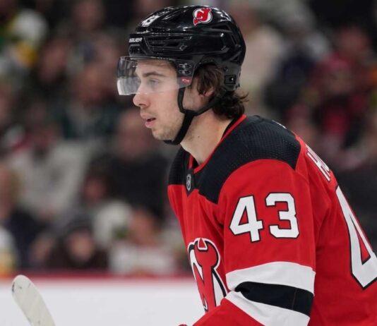 Quinn Hughes Trade Rumors: Minnesota Wild to New Jersey Devils? Close-up profile photograph of NHL star Quinn Hughes on the ice, wearing a New Jersey Devils home uniform and black helmet to visualize trade rumors that would reunite him with his brothers.