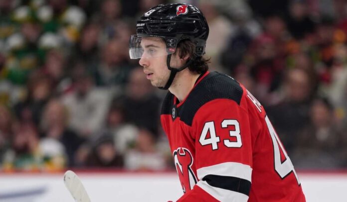 quinn-hughes-minnesota-wild-devils-trade Close-up profile photograph of NHL star Quinn Hughes on the ice, wearing a New Jersey Devils home uniform and black helmet to visualize trade rumors that would reunite him with his brothers.