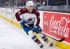 A photorealistic image of NHL forward Ross Colton, wearing his Colorado Avalanche away jersey (number 20 on sleeves and helmet), in an aggressive skating stride along the ice hockey boards. A large, prominent Coca-Cola advertising panel is clearly visible on the boards to his right.