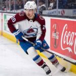 A photorealistic image of NHL forward Ross Colton, wearing his Colorado Avalanche away jersey (number 20 on sleeves and helmet), in an aggressive skating stride along the ice hockey boards. A large, prominent Coca-Cola advertising panel is clearly visible on the boards to his right.
