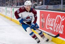 A photorealistic image of NHL forward Ross Colton, wearing his Colorado Avalanche away jersey (number 20 on sleeves and helmet), in an aggressive skating stride along the ice hockey boards. A large, prominent Coca-Cola advertising panel is clearly visible on the boards to his right.