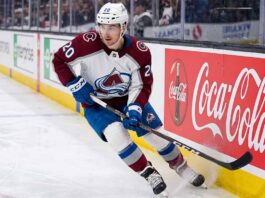 A photorealistic image of NHL forward Ross Colton, wearing his Colorado Avalanche away jersey (number 20 on sleeves and helmet), in an aggressive skating stride along the ice hockey boards. A large, prominent Coca-Cola advertising panel is clearly visible on the boards to his right.
