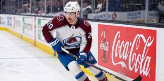 A photorealistic image of NHL forward Ross Colton, wearing his Colorado Avalanche away jersey (number 20 on sleeves and helmet), in an aggressive skating stride along the ice hockey boards. A large, prominent Coca-Cola advertising panel is clearly visible on the boards to his right.