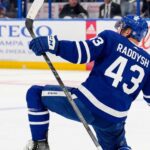 NHL defenseman Darren Raddysh wearing a Toronto Maple Leafs home uniform, kneeling on the ice in a celebratory pose holding his stick.
