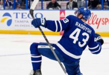 NHL defenseman Darren Raddysh wearing a Toronto Maple Leafs home uniform, kneeling on the ice in a celebratory pose holding his stick.