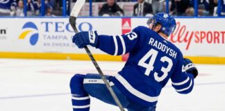 NHL defenseman Darren Raddysh wearing a Toronto Maple Leafs home uniform, kneeling on the ice in a celebratory pose holding his stick.