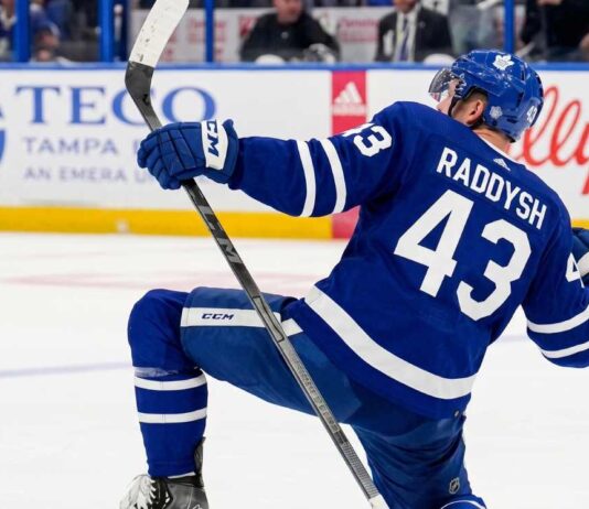 NHL defenseman Darren Raddysh wearing a Toronto Maple Leafs home uniform, kneeling on the ice in a celebratory pose holding his stick.