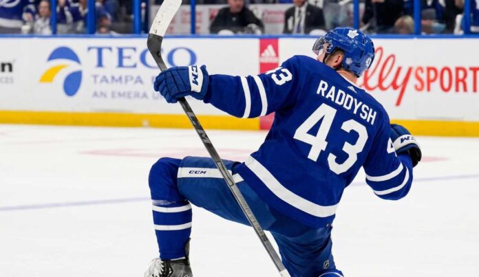 NHL defenseman Darren Raddysh wearing a Toronto Maple Leafs home uniform, kneeling on the ice in a celebratory pose holding his stick.
