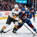 An intense photo capturing a puck battle between superstars Jack Eichel (#15) of the Vegas Golden Knights and Nathan MacKinnon (#29) of the Colorado Avalanche during Game 1 of the 2026 Western Conference Finals at Ball Arena. The background features the NHL Western Conference Finals sign and cheering fans.
