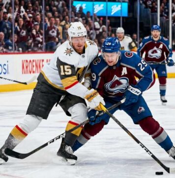 An intense photo capturing a puck battle between superstars Jack Eichel (#15) of the Vegas Golden Knights and Nathan MacKinnon (#29) of the Colorado Avalanche during Game 1 of the 2026 Western Conference Finals at Ball Arena. The background features the NHL Western Conference Finals sign and cheering fans.