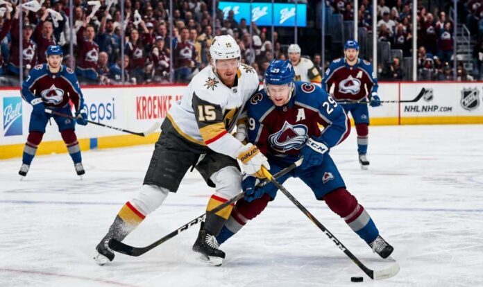 An intense photo capturing a puck battle between superstars Jack Eichel (#15) of the Vegas Golden Knights and Nathan MacKinnon (#29) of the Colorado Avalanche during Game 1 of the 2026 Western Conference Finals at Ball Arena. The background features the NHL Western Conference Finals sign and cheering fans.