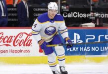 Buffalo Sabres defenseman Bowen Byram #4 skating with the puck on ice in a white away jersey, a top target in NHL trade rumors for teams like the Philadelphia Flyers and Chicago Blackhawks.