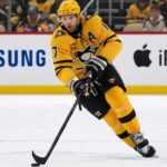 A photo of Pittsburgh Penguins forward and alternate captain Bryan Rust skating with the hockey puck in the team's yellow and black uniform during a game.