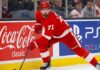 Detroit Red Wings forward Dylan Larkin (#71) in full action skating along the boards in an NHL arena, carrying the puck. Visible on the boards are prominent digital advertisements for Coca-Cola classic and PS5. Larkin is wearing his home red jersey and helmet. This image relates to the news surrounding the Dylan Larkin trade request from Detroit.