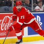 Detroit Red Wings forward Dylan Larkin (#71) in full action skating along the boards in an NHL arena, carrying the puck. Visible on the boards are prominent digital advertisements for Coca-Cola classic and PS5. Larkin is wearing his home red jersey and helmet. This image relates to the news surrounding the Dylan Larkin trade request from Detroit.