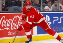Detroit Red Wings forward Dylan Larkin (#71) in full action skating along the boards in an NHL arena, carrying the puck. Visible on the boards are prominent digital advertisements for Coca-Cola classic and PS5. Larkin is wearing his home red jersey and helmet. This image relates to the news surrounding the Dylan Larkin trade request from Detroit.