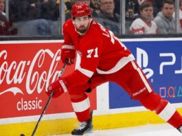 Detroit Red Wings forward Dylan Larkin (#71) in full action skating along the boards in an NHL arena, carrying the puck. Visible on the boards are prominent digital advertisements for Coca-Cola classic and PS5. Larkin is wearing his home red jersey and helmet. This image relates to the news surrounding the Dylan Larkin trade request from Detroit.