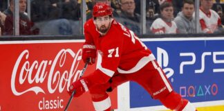 Dylan Larkin Trade Rumors: 4 Teams Targeting the Red Wings Captain Detroit Red Wings forward Dylan Larkin (#71) in full action skating along the boards in an NHL arena, carrying the puck. Visible on the boards are prominent digital advertisements for Coca-Cola classic and PS5. Larkin is wearing his home red jersey and helmet. This image relates to the news surrounding the Dylan Larkin trade request from Detroit.