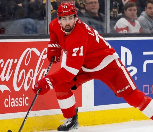 Detroit Red Wings forward Dylan Larkin (#71) in full action skating along the boards in an NHL arena, carrying the puck. Visible on the boards are prominent digital advertisements for Coca-Cola classic and PS5. Larkin is wearing his home red jersey and helmet. This image relates to the news surrounding the Dylan Larkin trade request from Detroit.