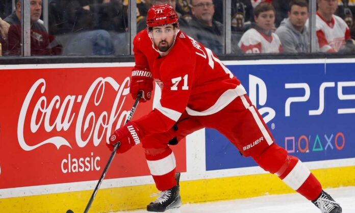 Detroit Red Wings forward Dylan Larkin (#71) in full action skating along the boards in an NHL arena, carrying the puck. Visible on the boards are prominent digital advertisements for Coca-Cola classic and PS5. Larkin is wearing his home red jersey and helmet. This image relates to the news surrounding the Dylan Larkin trade request from Detroit.
