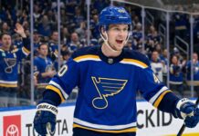 Elias Pettersson Trade Rumors: St. Louis Blues Linked to Vancouver Canucks Center Smiling Elias Pettersson wearing a St. Louis Blues home jersey with Blue Note logo and #40 helmet, celebrating on ice with cheering crowd; speculative trade visualization.