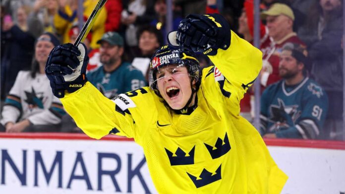 Team Sweden's Ivar Stenberg, wearing the iconic yellow and blue 'Tre Kronor' jersey with the number 15 and Svenska Spel logo, celebrating with a primal roar, raising his gloves and stick in triumph after scoring a goal during a hockey game, as San Jose Sharks fans look on in the background.