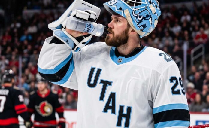 jacob-markstrom-utah-mammoth-devils-trade A close-up photograph of Jacob Markstrom, New Jersey Devils goaltender, wearing his white away jersey and custom mask while drinking from a water bottle on the ice, amidst trade rumors.