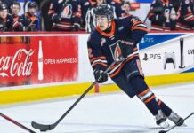 A detailed action photograph of hockey player JP Hurlbert, wearing number 22, competing for the Kamloops Blazers in their navy blue jersey with the flame 'B' logo. He is in full stride, stickhandling the puck on the ice. In the background, there are other Blazers players on the bench.