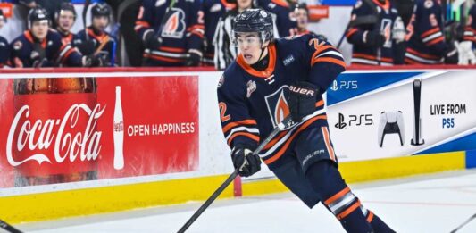 A detailed action photograph of hockey player JP Hurlbert, wearing number 22, competing for the Kamloops Blazers in their navy blue jersey with the flame 'B' logo. He is in full stride, stickhandling the puck on the ice. In the background, there are other Blazers players on the bench.