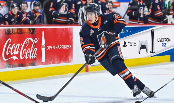 jp-hurlbert-los-angeles-kings-2026-draft A detailed action photograph of hockey player JP Hurlbert, wearing number 22, competing for the Kamloops Blazers in their navy blue jersey with the flame 'B' logo. He is in full stride, stickhandling the puck on the ice. In the background, there are other Blazers players on the bench.
