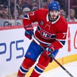 Montreal Canadiens Trade Rumors: Will Kirby Dach Be Traded? Montreal Canadiens center Kirby Dach #77 skating on the ice during an NHL game, with the PS5 and Coca-Cola logos visible on the arena boards behind him. This image relates to current trade rumors surrounding Dach.
