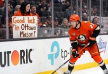 Anaheim Ducks forward Mason McTavish skating along the boards while a fan in the stands holds a sign reading "Good luck in Ottawa Mason!" amidst Ottawa Senators trade rumors.