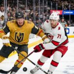 Action shot of Vegas Golden Knights forward Jack Eichel and a Carolina Hurricanes opponent battling for the puck on the ice during Game 1 of the 2026 Stanley Cup Final, with cheering fans, NHL logos, and the 'Vegas Golden Knights' rink board in the background.