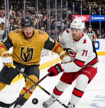 Action shot of Vegas Golden Knights forward Jack Eichel and a Carolina Hurricanes opponent battling for the puck on the ice during Game 1 of the 2026 Stanley Cup Final, with cheering fans, NHL logos, and the 'Vegas Golden Knights' rink board in the background.