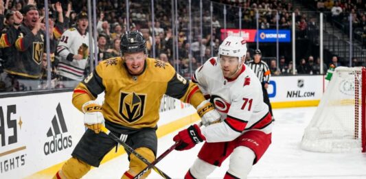 Action shot of Vegas Golden Knights forward Jack Eichel and a Carolina Hurricanes opponent battling for the puck on the ice during Game 1 of the 2026 Stanley Cup Final, with cheering fans, NHL logos, and the 'Vegas Golden Knights' rink board in the background.