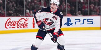 Zach Werenski, Columbus Blue Jackets star defenseman, during an NHL game, seen in his white away jersey. The photo captures him looking ahead, the primary focus of growing trade speculation for the team.
