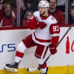 Alex DeBrincat Point Projections 2026-27: Detroit Red Wings Fantasy Outlook Alex DeBrincat celebrating in a Detroit Red Wings jersey beside PS5 and Coca-Cola board ads for his 2026-27 fantasy hockey point projection.