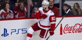 Alex DeBrincat Point Projections 2026-27: Detroit Red Wings Fantasy Outlook Alex DeBrincat celebrating in a Detroit Red Wings jersey beside PS5 and Coca-Cola board ads for his 2026-27 fantasy hockey point projection.