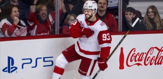 Alex DeBrincat Point Projections 2026-27: Detroit Red Wings Fantasy Outlook Alex DeBrincat celebrating in a Detroit Red Wings jersey beside PS5 and Coca-Cola board ads for his 2026-27 fantasy hockey point projection.