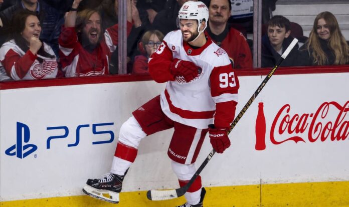 alex-debrincat-detroit-red-wings-fantasy-hockey-projection-2026-27 Alex DeBrincat celebrating in a Detroit Red Wings jersey beside PS5 and Coca-Cola board ads for his 2026-27 fantasy hockey point projection.