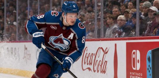 Cale Makar Point Projections 2026-27: Can He Return to 90 Points? Cale Makar skating for the Colorado Avalanche as a right-shot defenseman beside Coca-Cola and Nintendo Switch board ads during a 2026-27 Fantasy Hockey projection.