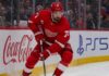 Dylan Larkin’s Three-Team List Has Trapped the Detroit Red Wings Dylan Larkin skating in a Detroit Red Wings home jersey beside Coca-Cola and PS5 rink-board ads amid NHL trade rumors.