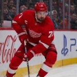 Dylan Larkin’s Three-Team List Has Trapped the Detroit Red Wings Dylan Larkin skating in a Detroit Red Wings home jersey beside Coca-Cola and PS5 rink-board ads amid NHL trade rumors.