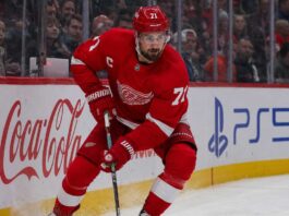 Dylan Larkin’s Three-Team List Has Trapped the Detroit Red Wings Dylan Larkin skating in a Detroit Red Wings home jersey beside Coca-Cola and PS5 rink-board ads amid NHL trade rumors.
