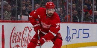 Dylan Larkin skating in a Detroit Red Wings home jersey beside Coca-Cola and PS5 rink-board ads amid NHL trade rumors.