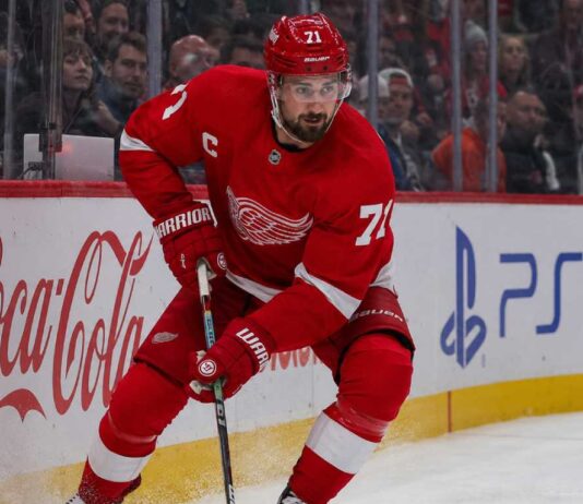 Dylan Larkin’s Three-Team List Has Trapped the Detroit Red Wings Dylan Larkin skating in a Detroit Red Wings home jersey beside Coca-Cola and PS5 rink-board ads amid NHL trade rumors.