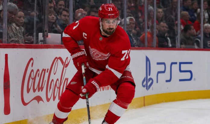 dylan-larkin-detroit-red-wings-trade-rumors Dylan Larkin skating in a Detroit Red Wings home jersey beside Coca-Cola and PS5 rink-board ads amid NHL trade rumors.