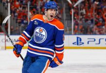 Evan Bouchard Point Projections 2026-27: Edmonton Oilers Forecast Evan Bouchard celebrates in an Edmonton Oilers home jersey beside a PS5 rink-board ad ahead of his 2026-27 fantasy hockey projection