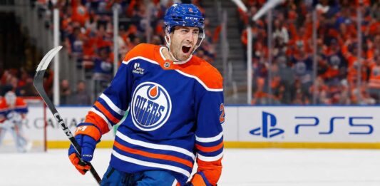 Evan Bouchard Point Projections 2026-27: Edmonton Oilers Forecast Evan Bouchard celebrates in an Edmonton Oilers home jersey beside a PS5 rink-board ad ahead of his 2026-27 fantasy hockey projection