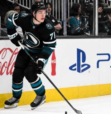 Macklin Celebrini Point Projections 2026-27: San Jose Sharks Fantasy Hockey Outlook Macklin Celebrini skating for the San Jose Sharks with Coca-Cola and PS5 board ads, highlighting his 2026-27 fantasy hockey point projection